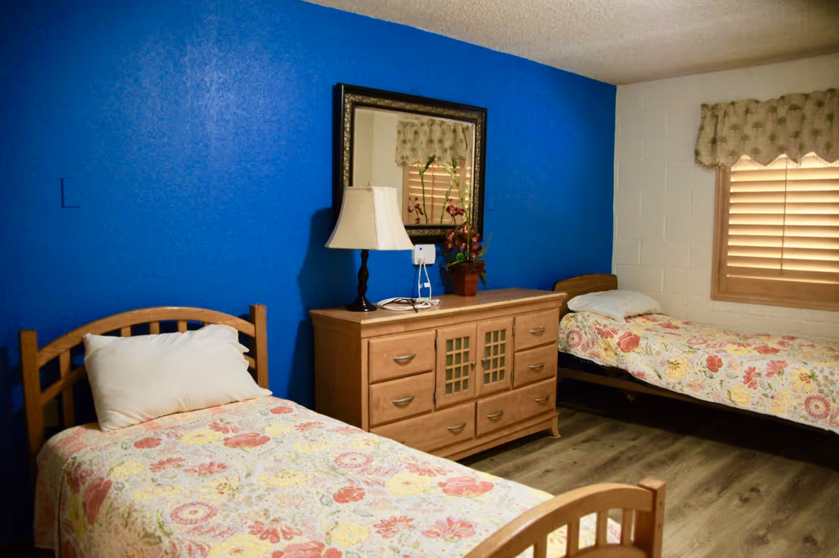 A bedroom with two single beds featuring floral bedspreads and white pillows. Between the beds is a wooden dresser with multiple drawers, a table lamp, a potted plant, and a framed mirror hanging above it. The wall behind the dresser is painted bright blue, while the adjacent wall is white with a window covered by wooden blinds and a floral valance. The floor is wood laminate.