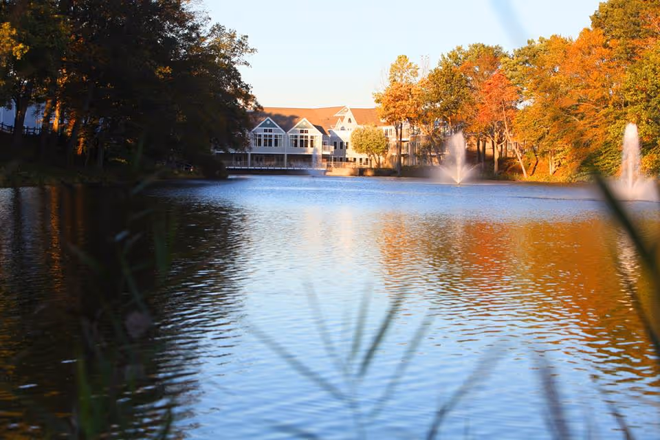A lakeside view of a senior living facility building with fountains and autumn trees reflected in the water.