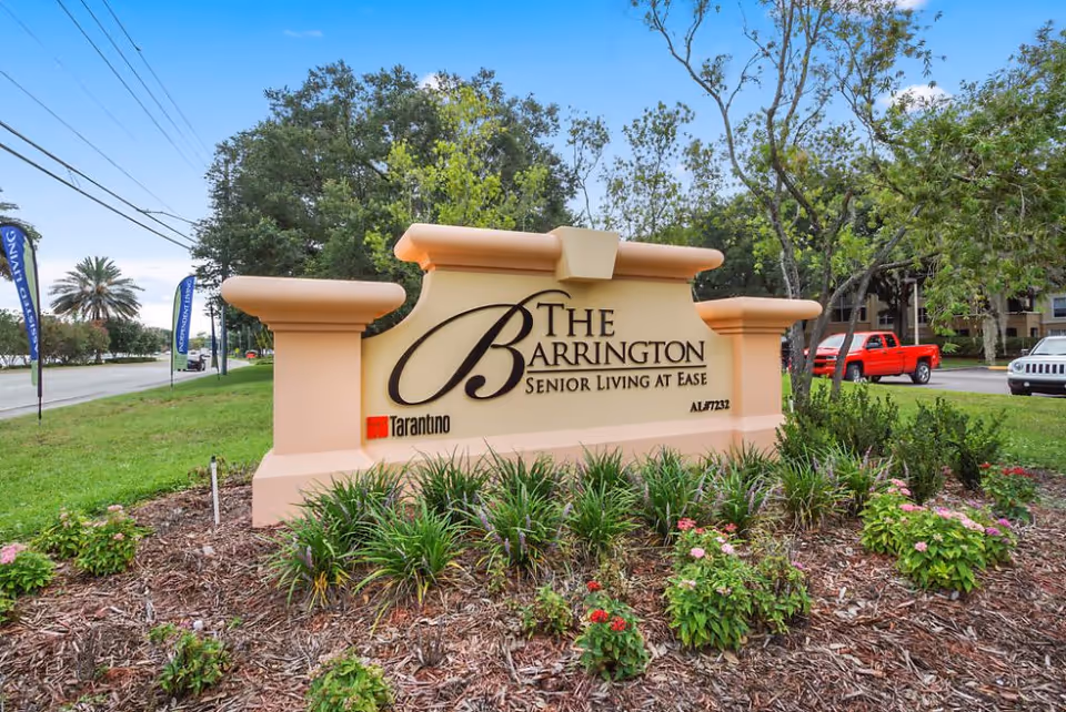 Outdoor view of a large beige sign with decorative columns on each side, displaying the name 'The Barrington Senior Living at Ease' surrounded by landscaped greenery and flowers, with a road and trees in the background.
