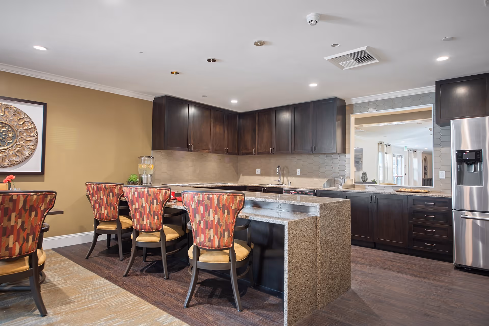 A modern kitchen and dining area with dark wood cabinets, a granite countertop island, and stainless steel refrigerator. The dining area features a table with six chairs upholstered in a red and beige patterned fabric. The walls are painted beige, and there is a decorative round wall art piece on one wall. The kitchen has a window opening to an adjacent room.