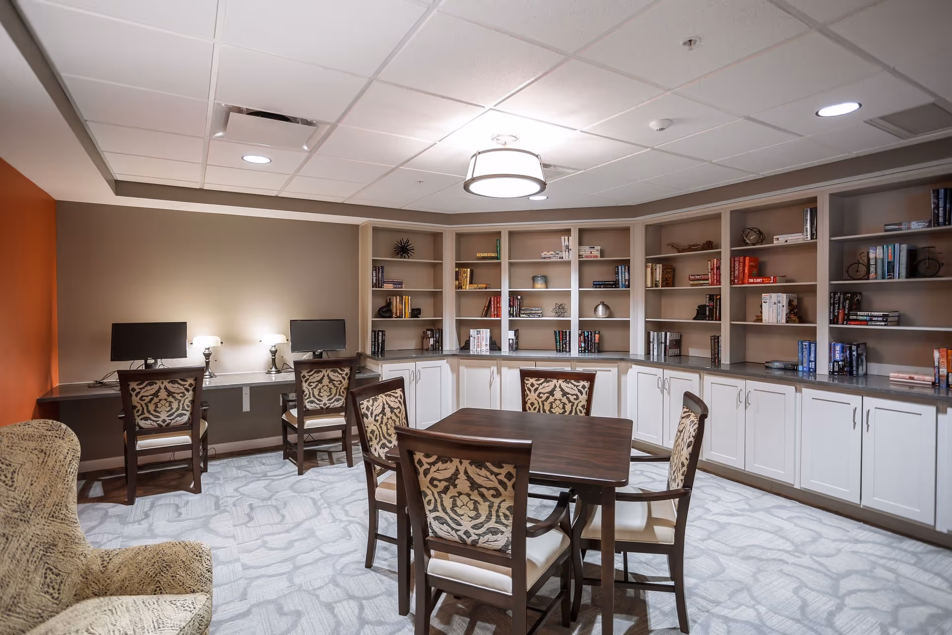 A cozy senior living facility common room with a square wooden table surrounded by four patterned chairs in the center. Along the back wall are built-in bookshelves filled with books and decorative items, with a countertop below. Two computer stations with monitors and lamps are set up against the left wall. The room has a patterned carpet, neutral wall colors with one orange accent wall, and a ceiling light fixture.