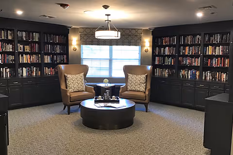 Cozy library-style sitting area with floor-to-ceiling bookshelves, two armchairs facing a round coffee table beneath a central light and a window.