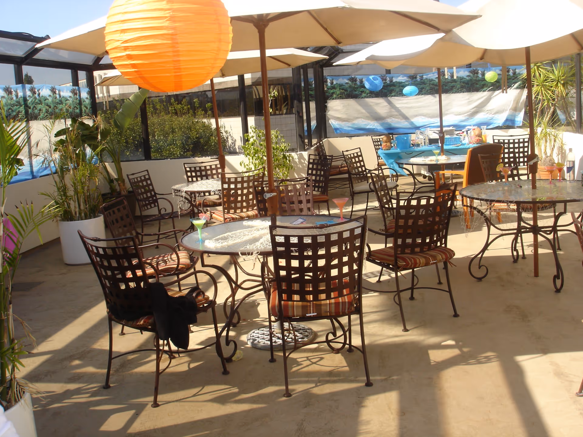 Outdoor patio area with multiple round glass tables and metal chairs with striped cushions. Large beige umbrellas provide shade over the tables. There are potted plants around the patio and colorful paper lanterns hanging above. A table in the background is covered with a blue tablecloth and various items on top.
