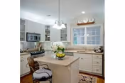Bright kitchen with white cabinetry, a central island with a bowl of lemons and a vase of white flowers, a stainless steel microwave and stove, a window above the sink, and a cushioned chair at the island.