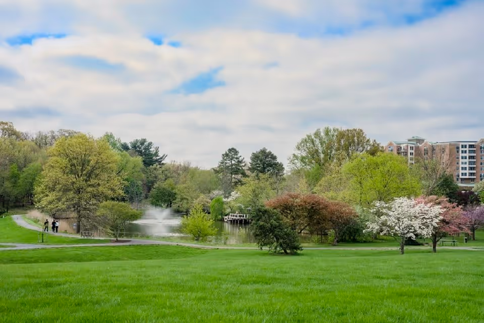 A scenic outdoor park area with lush green grass, various trees in bloom, a pond with a water fountain, and a walking path where two people are strolling. In the background, there is a multi-story building partially visible behind the trees under a partly cloudy sky.