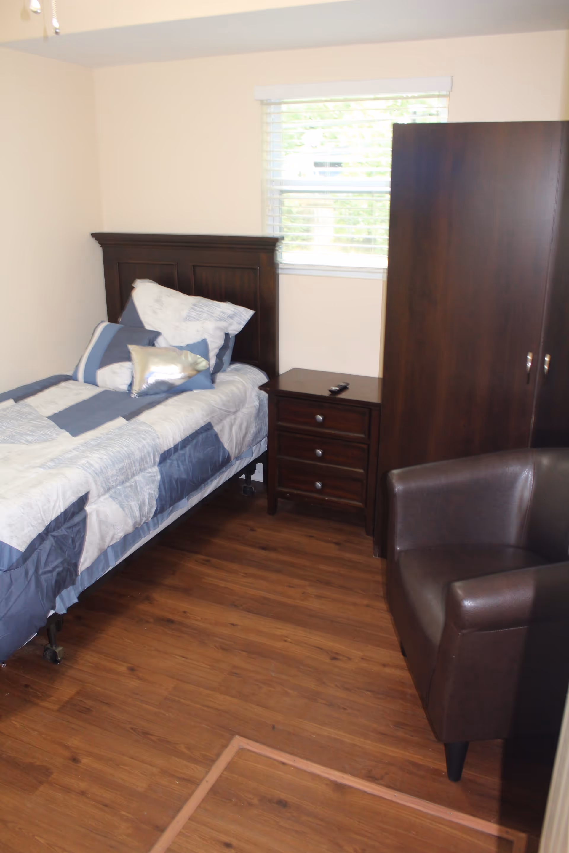A small bedroom with a single bed featuring a dark wooden headboard and blue and white bedding. Next to the bed is a dark wooden nightstand with three drawers and a remote control on top. There is a window with white blinds above the nightstand. To the right of the nightstand is a dark wooden wardrobe. In the foreground, there is a dark brown leather armchair. The floor is wooden.