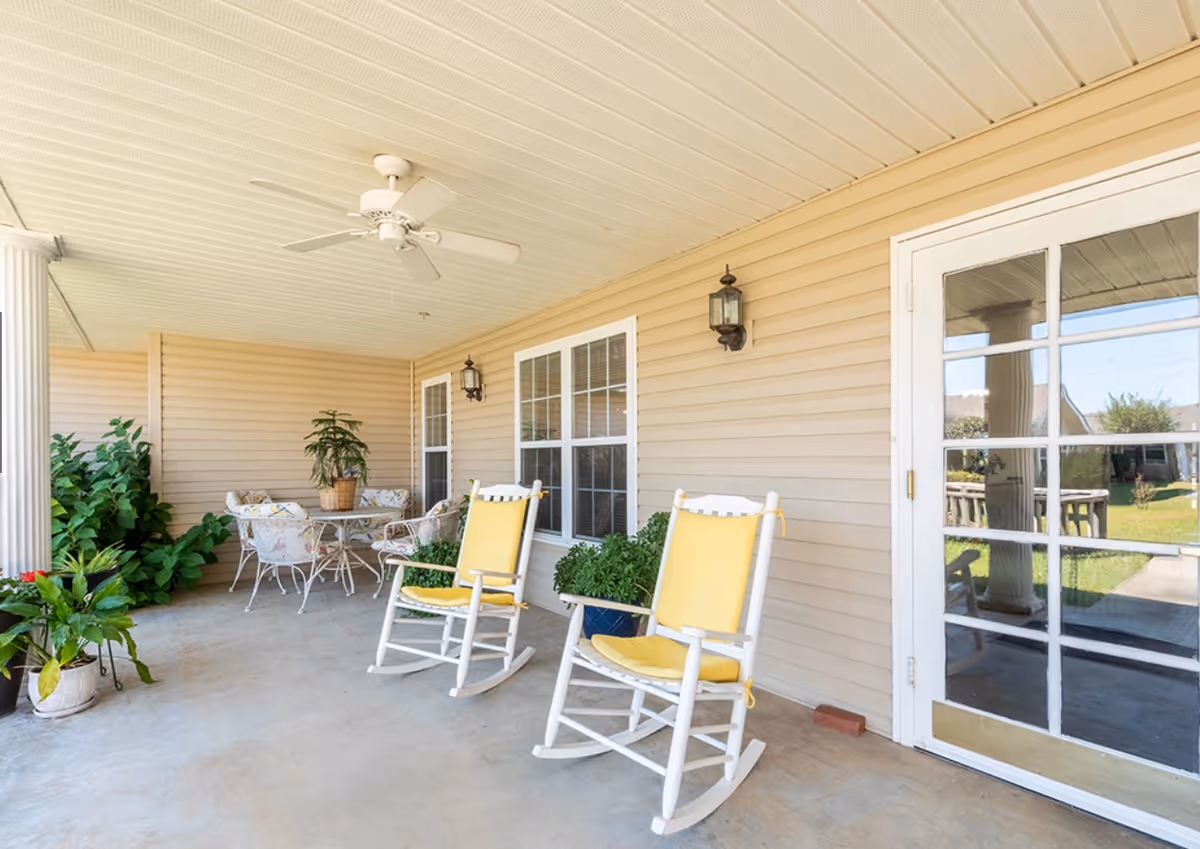 Covered outdoor patio area with two white rocking chairs featuring yellow cushions, a round table with four cushioned chairs, several potted plants, beige siding walls, two wall-mounted lantern lights, a white ceiling fan, and a glass door leading inside.