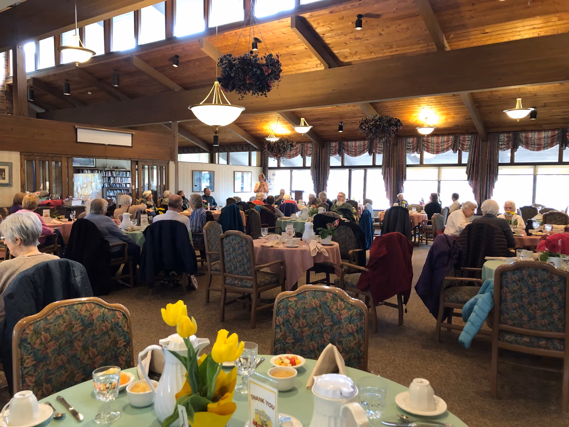 A dining room in a senior living facility with multiple round tables covered with pastel-colored tablecloths. Elderly residents are seated around the tables, engaged in conversation and dining. The room has a high wooden ceiling with hanging light fixtures and floral decorations. Large windows with curtains allow natural light to fill the space.