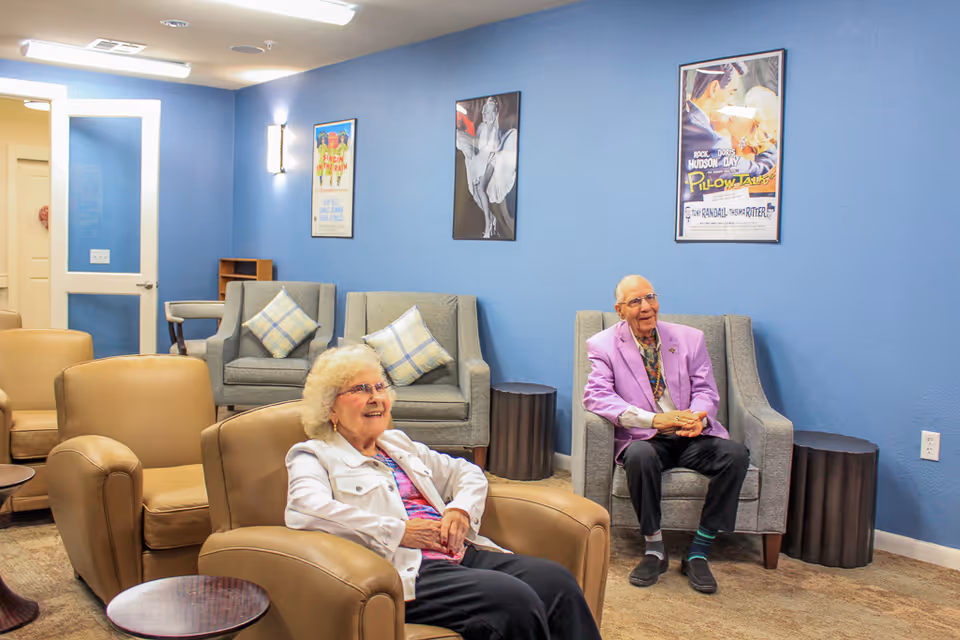 Two elderly individuals sitting and smiling in a comfortable lounge area with blue walls, armchairs, and framed vintage movie posters on the wall.