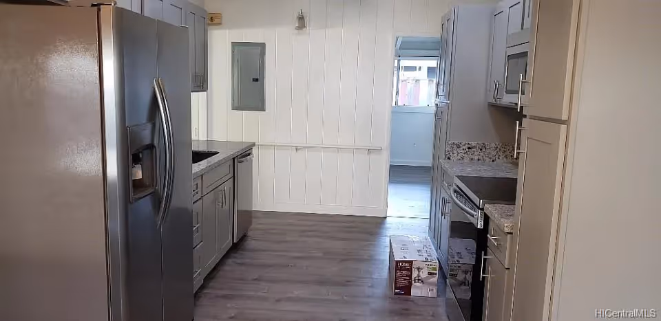 Modern kitchen with gray cabinets, stainless steel refrigerator, dishwasher, oven, and microwave. The kitchen has wood flooring and white paneled walls, with a window at the far end letting in natural light. There is a box on the floor near the oven.