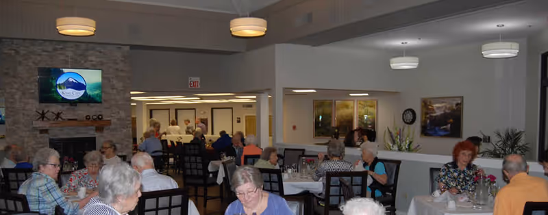A group of older adults seated at tables eating and socializing in a bright senior living dining room with a stone fireplace and mounted TV.
