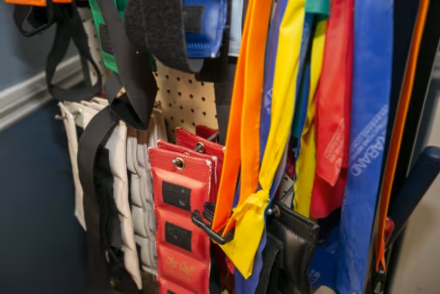 Close-up view of various colorful exercise resistance bands and wrist or ankle weights hanging on hooks against a pegboard wall in a room with blue walls.