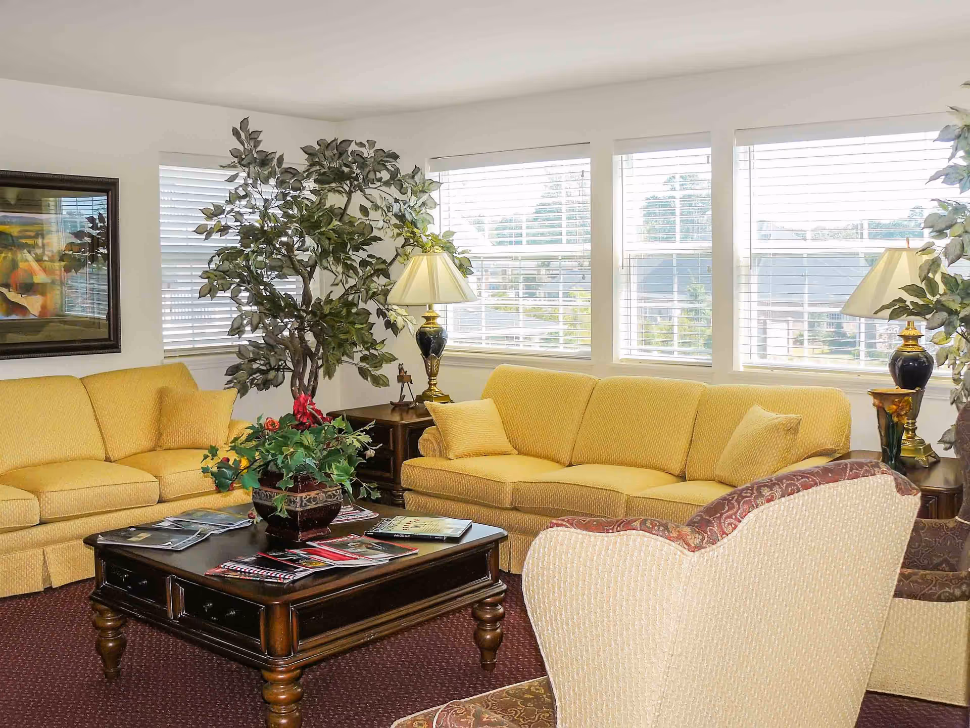 Sunlit living room with yellow sofas, armchairs, a wooden coffee table, lamps and large windows.