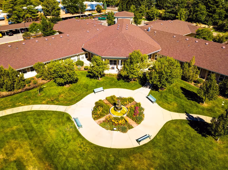 Aerial view of a senior living facility with a large building featuring a red roof surrounded by green lawns, trees, and a circular walkway with benches and a central fountain with flowers.
