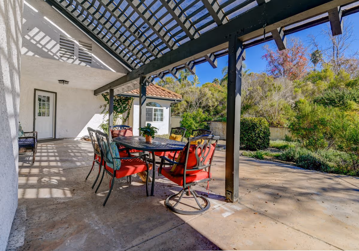 Outdoor patio area with a pergola overhead casting shadows on the concrete floor. There is a black metal table with a mosaic top surrounded by six chairs with red cushions. The patio is adjacent to a white building with a door and window, and there is greenery and trees in the background under a clear blue sky.