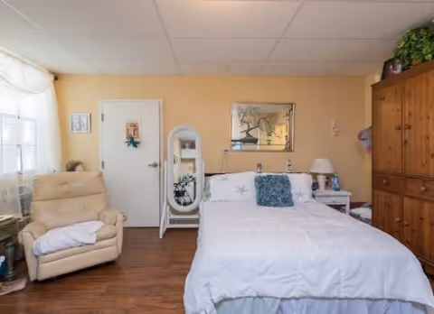 A cozy bedroom with a white bedspread on a bed, a blue decorative pillow, a wooden armoire on the right, a beige recliner chair on the left, a white standing mirror, and a window with sheer curtains letting in natural light.
