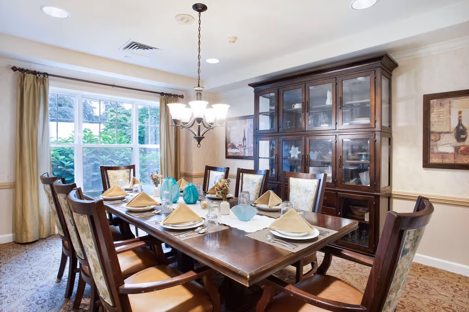 A well-lit dining room with a large wooden dining table set for eight people. Each place setting includes a plate, napkin, glass, and silverware. The room features a large window with beige curtains, a chandelier hanging above the table, a wooden china cabinet filled with dishes, and framed artwork on the walls.