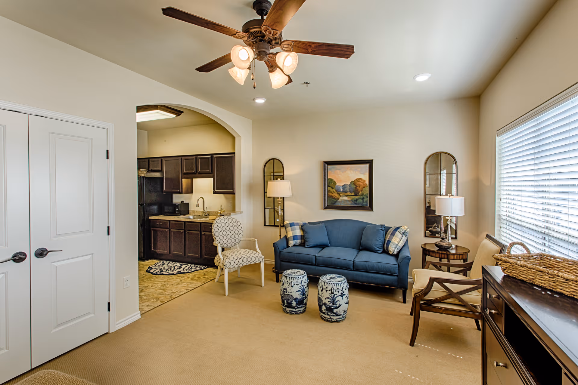A cozy living room area with a blue sofa, two patterned stools, a white armchair, and a wooden chair. There is a ceiling fan with lights, a painting on the wall, two mirrors, and a lamp. The room has beige walls and carpet, with a window covered by blinds letting in natural light. An open archway leads to a kitchen with dark cabinets and a black refrigerator.
