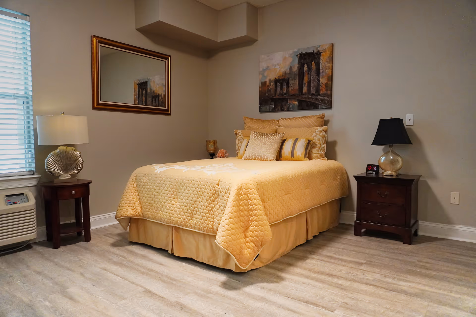 A neatly made bed with yellow quilt and decorative pillows between two nightstands in a softly lit bedroom with wall art.