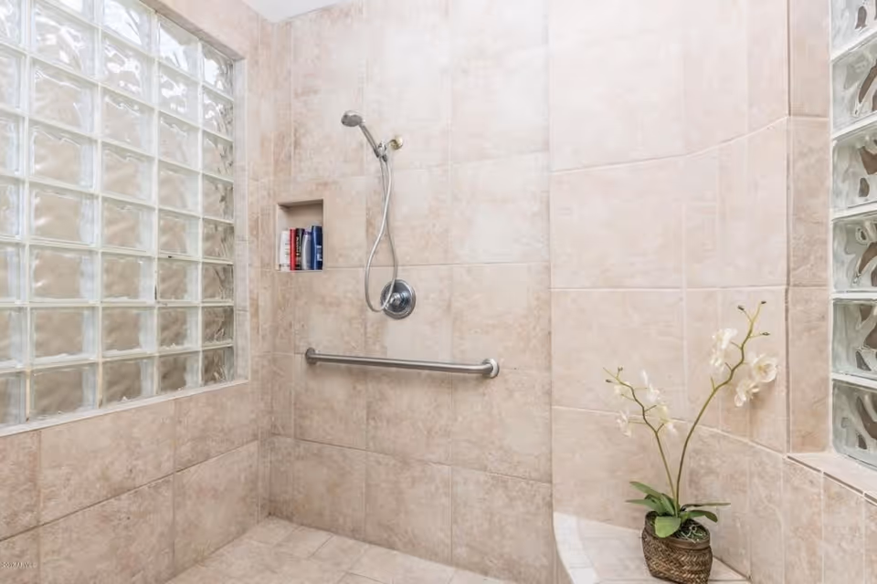 A tiled walk-in shower with beige tiles covering the walls and floor. The shower features a handheld showerhead, a built-in niche holding shampoo bottles, a horizontal grab bar, and a glass block window allowing natural light. There is a small bench with a potted white orchid plant placed on it.