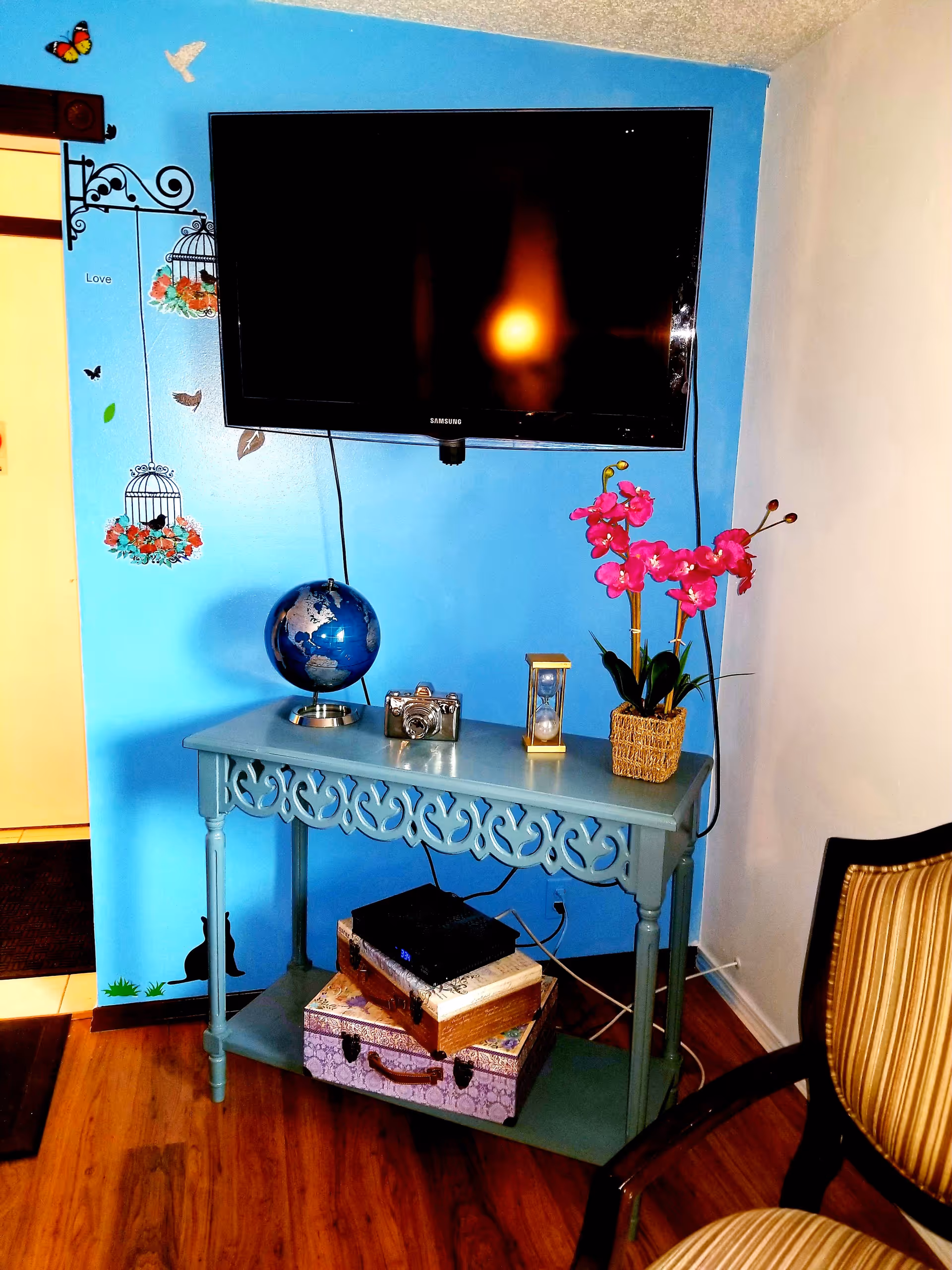 A corner of a room with a mounted flat-screen Samsung TV on a blue wall decorated with birdcage and butterfly wall decals. Below the TV is a decorative blue table holding a globe, a vintage camera, an hourglass, and a potted pink orchid. Under the table are two decorative suitcases and a black electronic device. Part of a striped upholstered chair is visible on the right side.