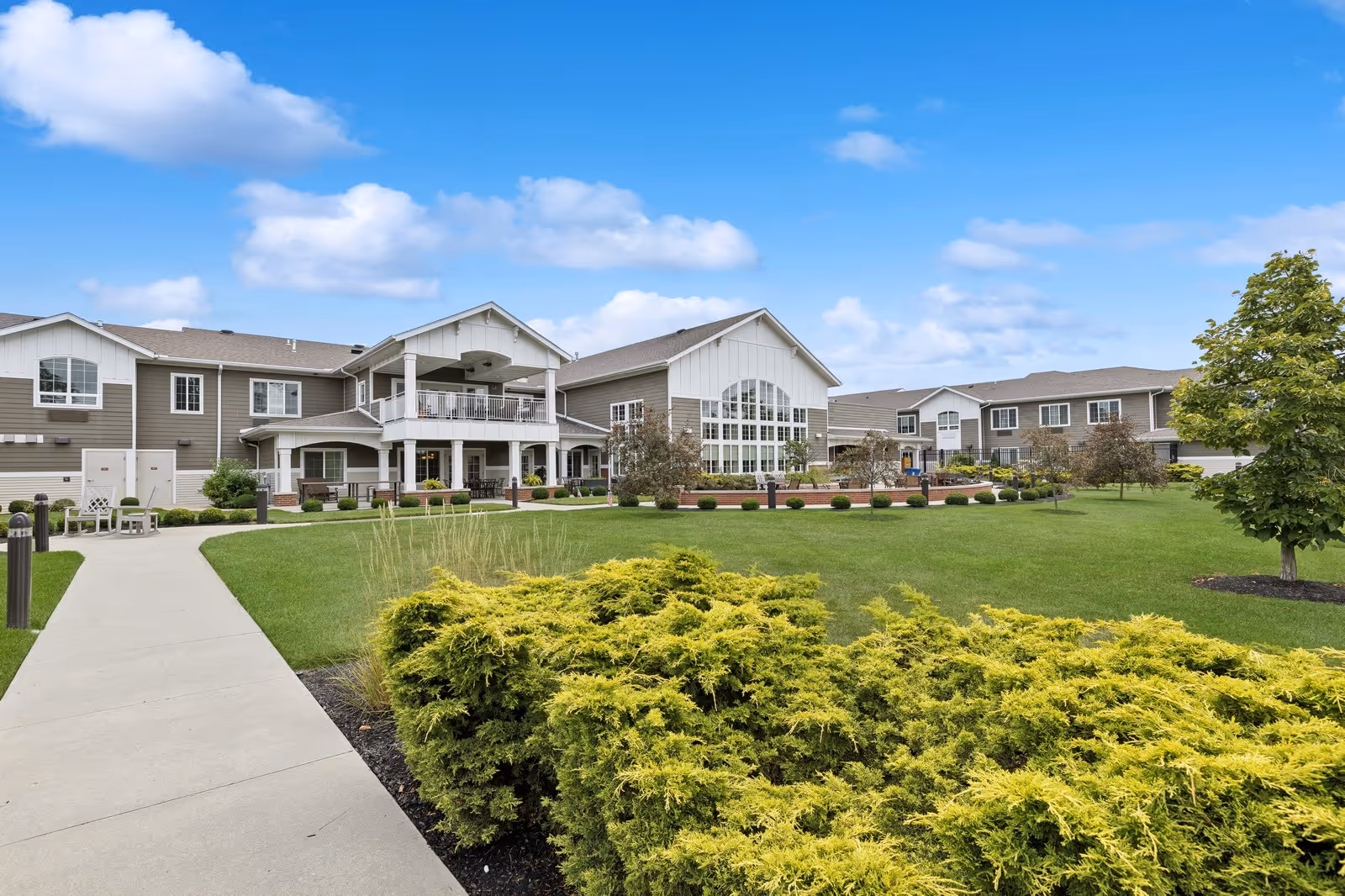 Exterior view of The Enclave of Springboro senior living facility with a well-maintained lawn, shrubs, trees, and a paved walkway leading to the building under a partly cloudy blue sky.