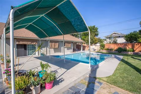Backyard with a covered patio, swimming pool, potted plants and lawn beside a single-story house.