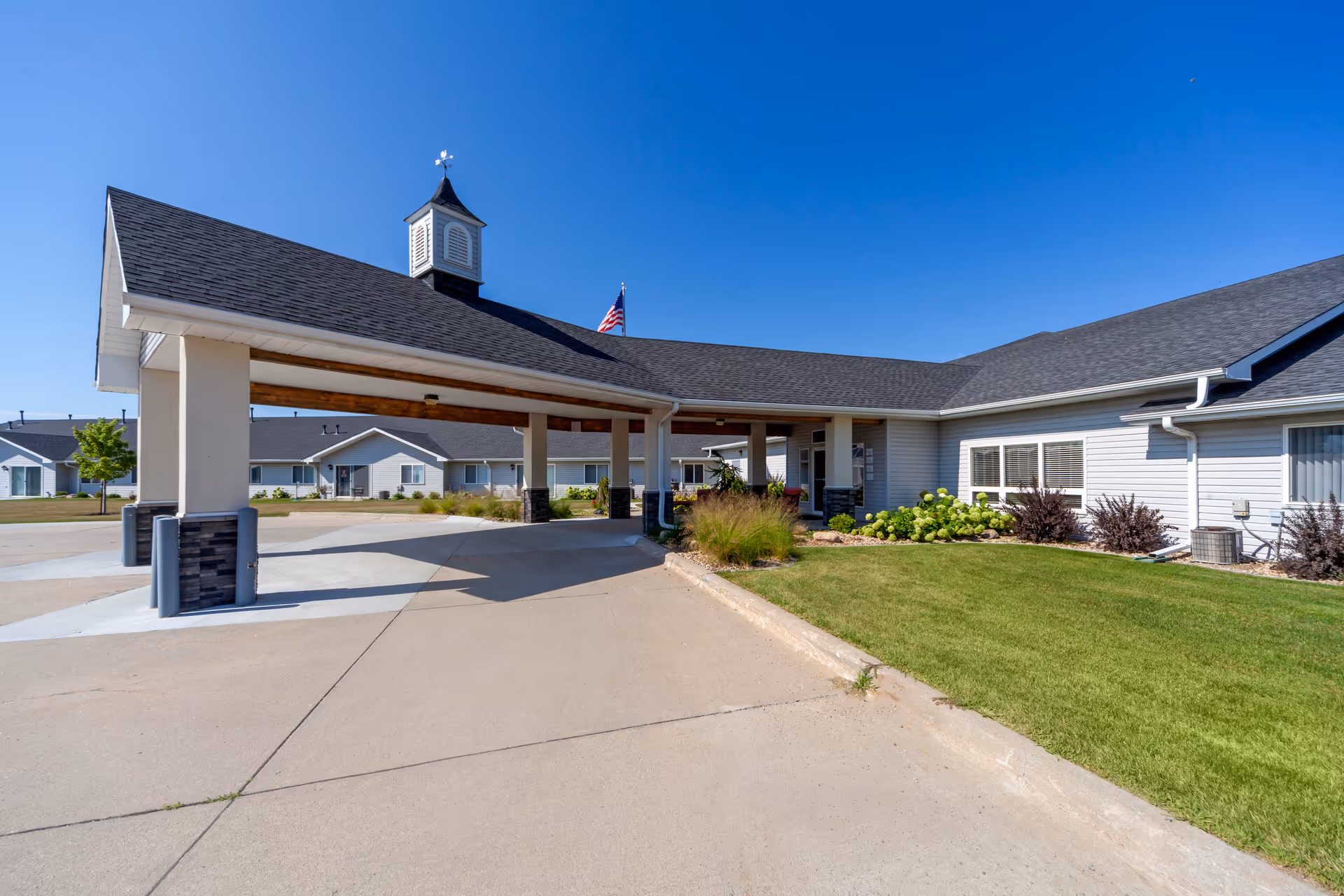 Exterior view of Kinship Pointe at Wayne facility showing a covered entrance with a cupola on the roof, an American flag, well-maintained lawn, and multiple single-story buildings under a clear blue sky.