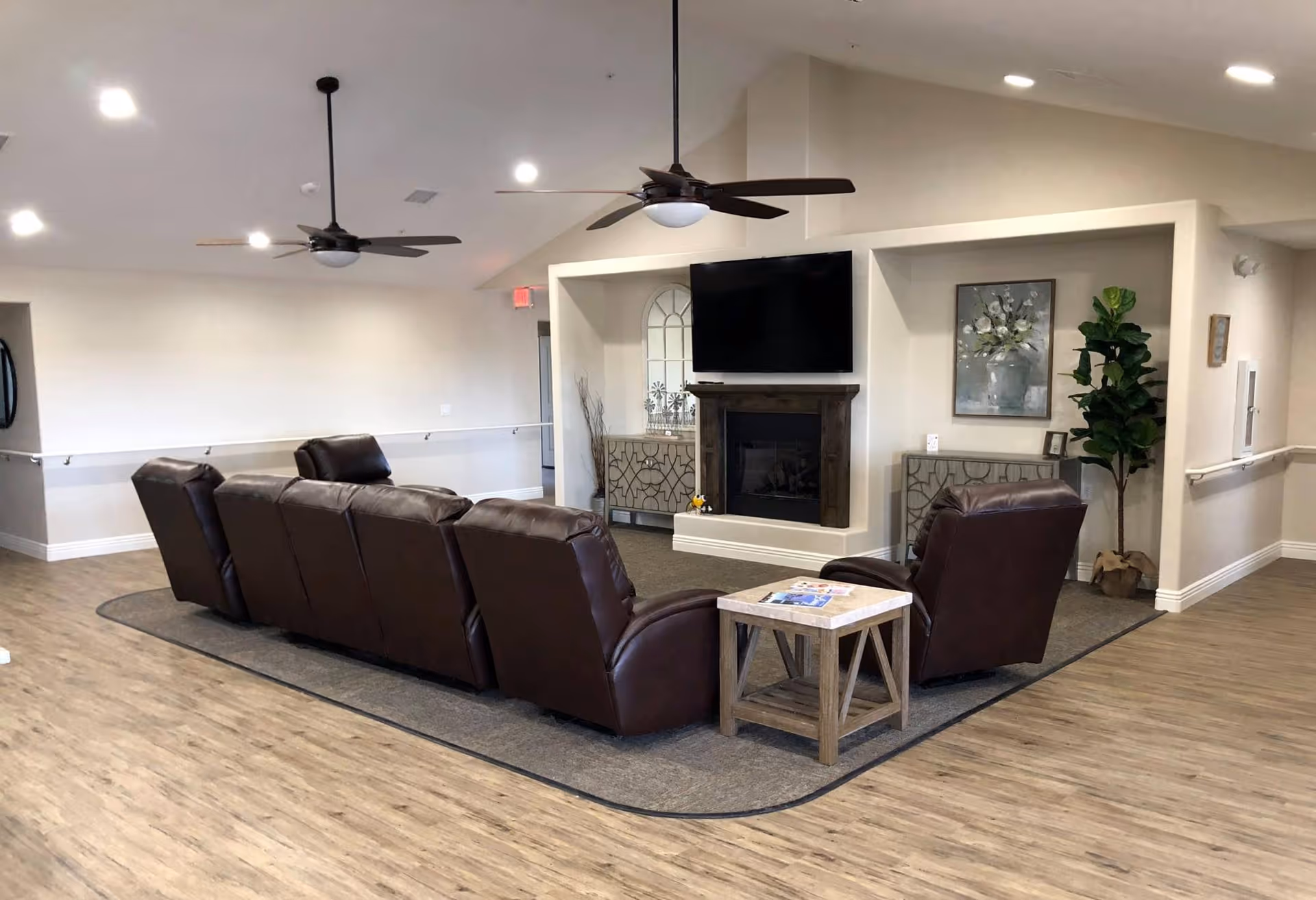 Spacious common living room with a row of leather recliners and chairs facing a TV mounted above a fireplace, with side tables, plants, and ceiling fans.