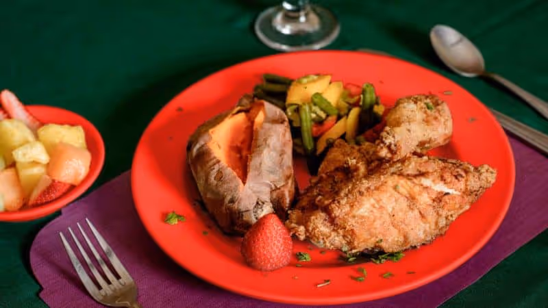 A red plate with a baked sweet potato, fried chicken drumstick and thigh, steamed mixed vegetables, and a strawberry on a green tablecloth with a purple napkin. A small bowl of mixed fruit and silverware are also visible.
