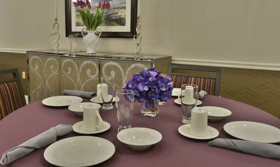 A round dining table set with white plates, cups, bowls, glasses, and gray napkins on a purple tablecloth. A small vase with purple flowers is in the center. In the background, there is a decorative sideboard with a white vase holding pink tulips and a framed painting on the wall.