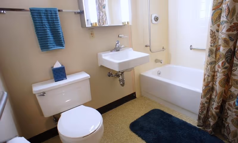 A clean bathroom featuring a white toilet with a blue tissue box on top, a white wall-mounted sink with a faucet, a bathtub with a floral-patterned shower curtain, a blue bath mat on the floor, and a blue towel hanging on a towel rack.