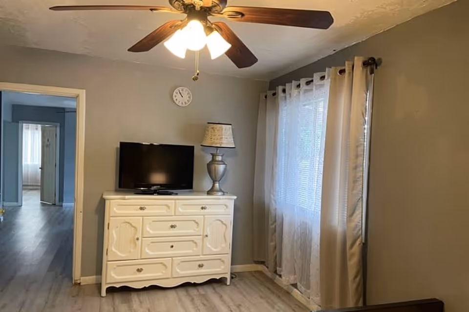 A cozy room with light gray walls and wood flooring featuring a white dresser with a flat-screen TV and a decorative lamp on top. A ceiling fan with lights is mounted on the ceiling. There is a window with sheer and solid curtains on the right side, and an open doorway leading to another room on the left.