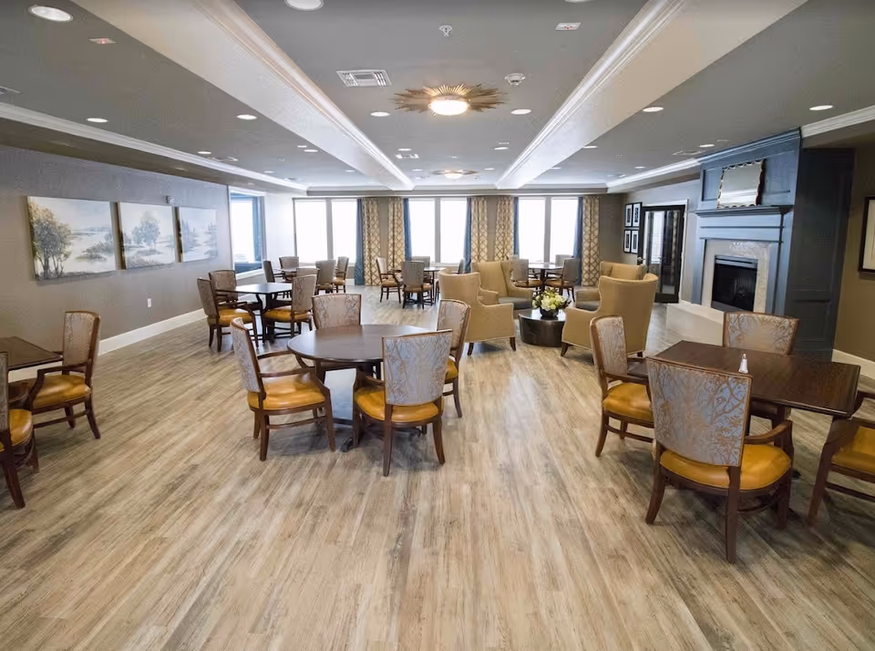 A spacious senior living common area with multiple round and square tables surrounded by chairs, a seating area with armchairs around a coffee table, large windows with patterned curtains, a fireplace with a decorative mirror above it, and three landscape paintings on one wall.