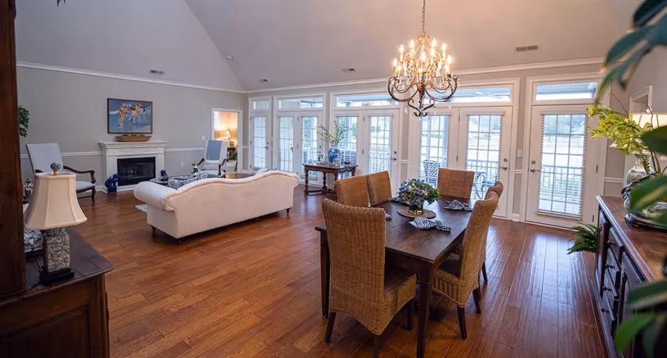 Open-plan living and dining area with a wooden table and wicker chairs, chandelier, white sofa, fireplace, and wall of glass doors.