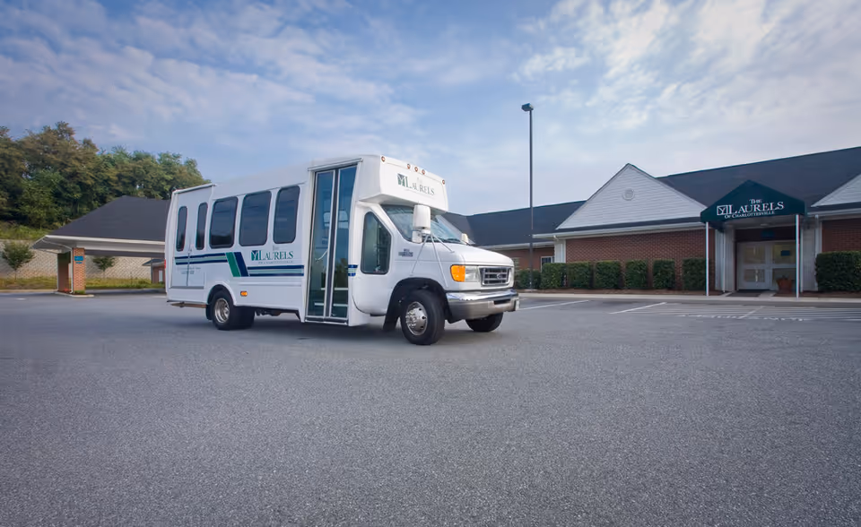A white shuttle bus with The Laurels of Charlottesville logo parked in an empty parking lot in front of a single-story brick building with a green awning displaying The Laurels of Charlottesville name.