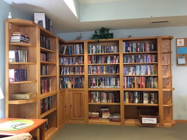 Wooden bookshelves filled with books line a wall in a small indoor reading area with a table visible in the foreground.