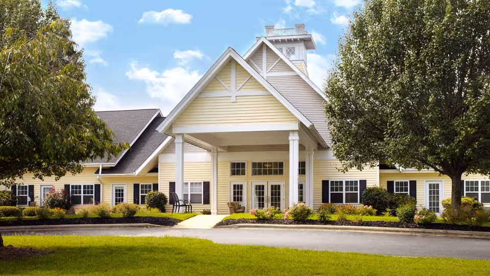 Front exterior view of a senior living facility building with a covered entrance, beige siding, white trim, black shutters, and surrounded by green trees and landscaping under a partly cloudy sky.