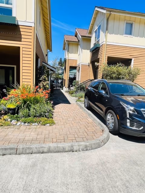 View of a paved walkway between two buildings with beige and brown siding under a clear blue sky. There are plants and flowers along the left side of the walkway, and a black SUV is parked on the right side near the building.
