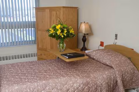 A senior living facility bedroom with a bed, wooden cabinet, lamp, and vase of yellow flowers.