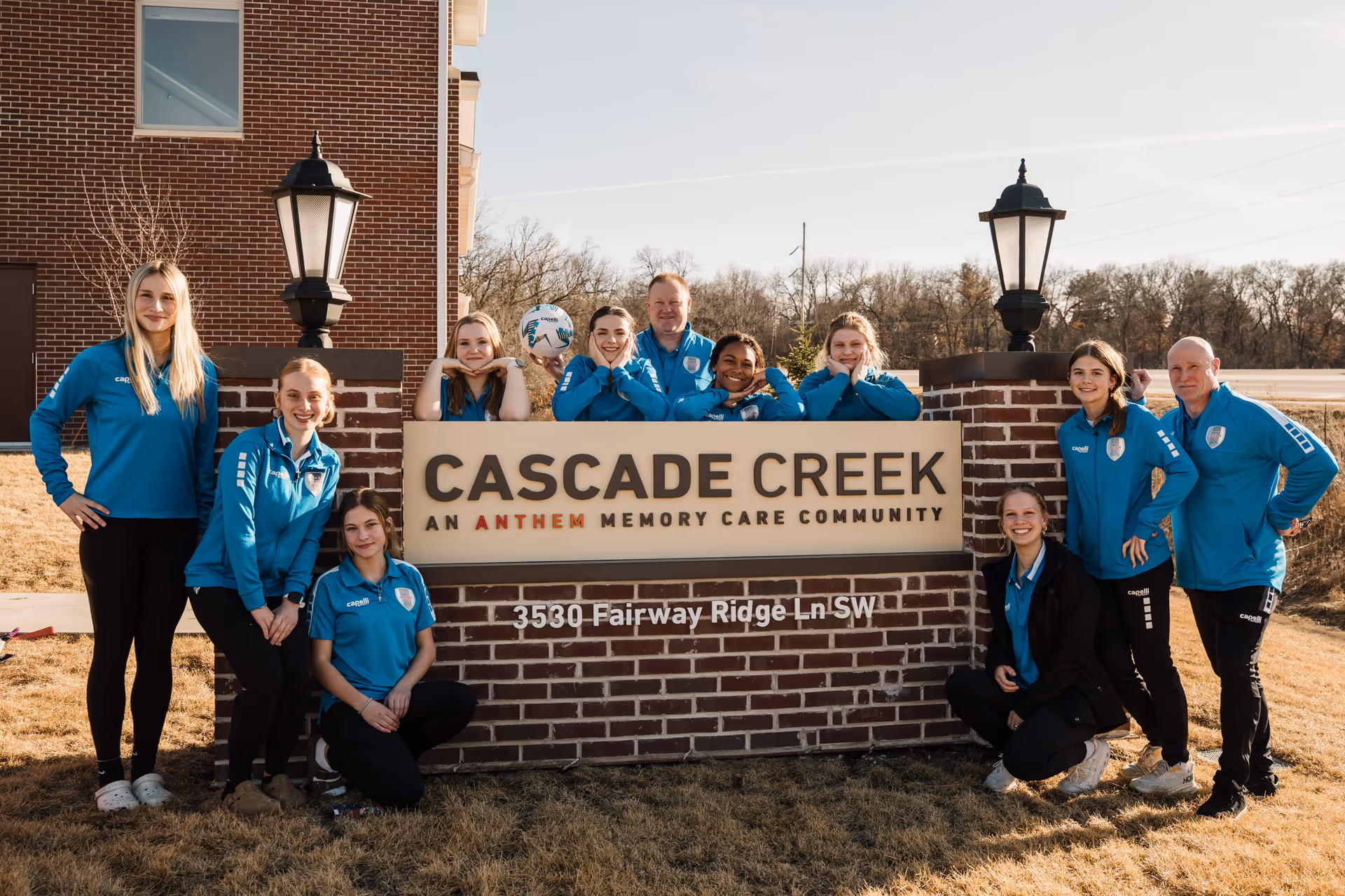 Staff in blue jackets pose around a brick sign that reads 'CASCADE CREEK, AN ANTHEM MEMORY CARE COMMUNITY' outside the facility.