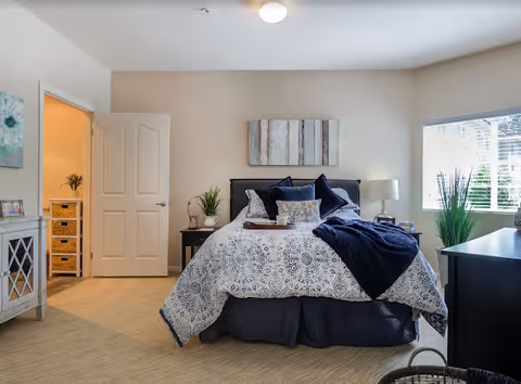 Sunlit furnished bedroom with a made bed, nightstands, plants, and a window.