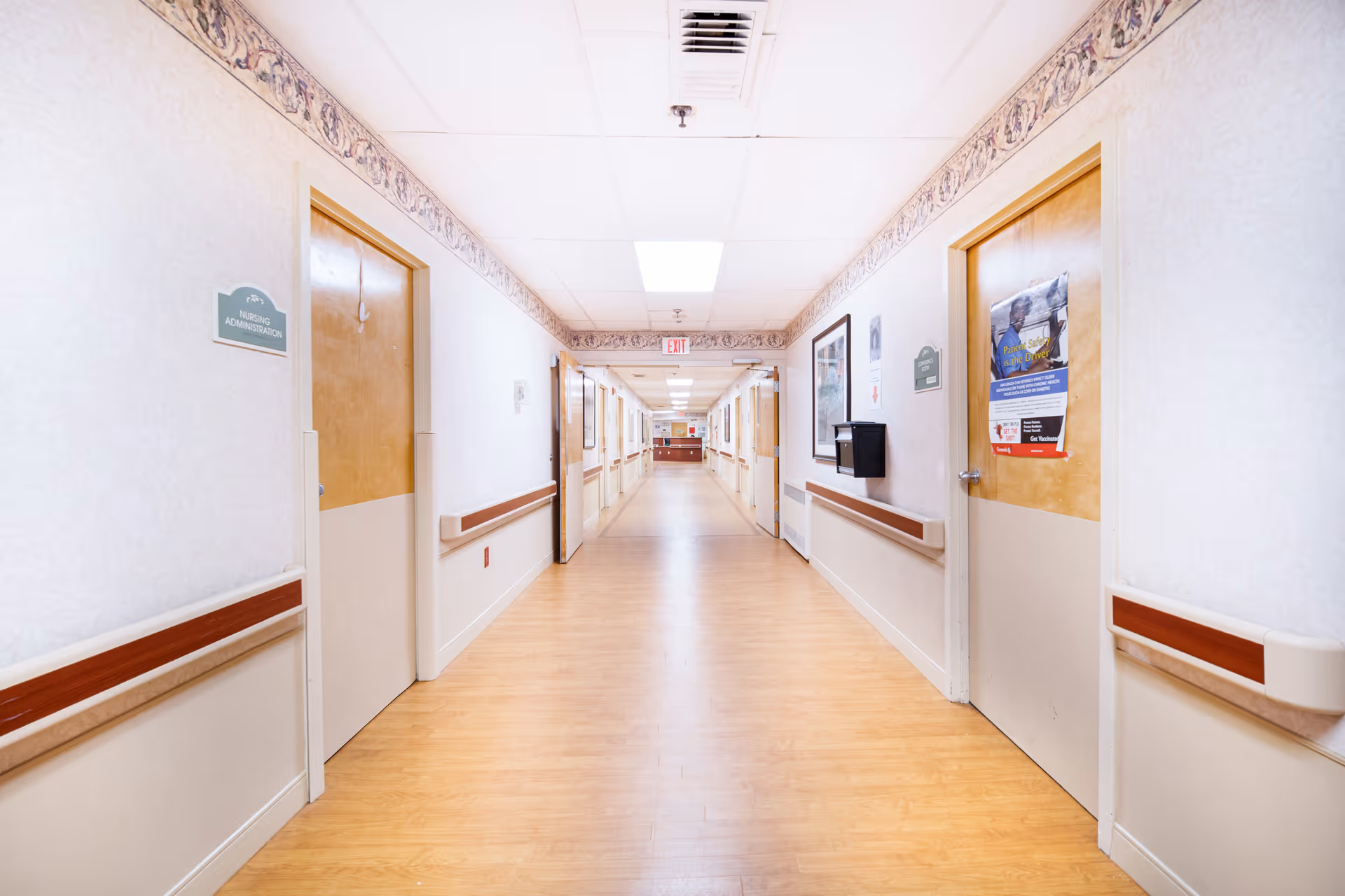A long, well-lit hallway in a senior living facility with light wood flooring, white walls, and wooden doors on both sides. The hallway has handrails along the walls and decorative border trim near the ceiling. Signs on the doors indicate rooms such as Nursing Administration. There are framed pictures and a bulletin board on the walls, and an exit sign is visible at the far end.