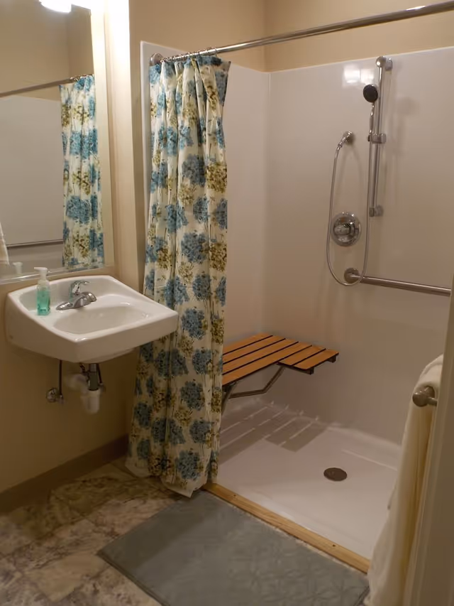 Accessible bathroom with a wall-mounted sink, floral shower curtain, fold-down wooden shower bench, and handheld showerhead.