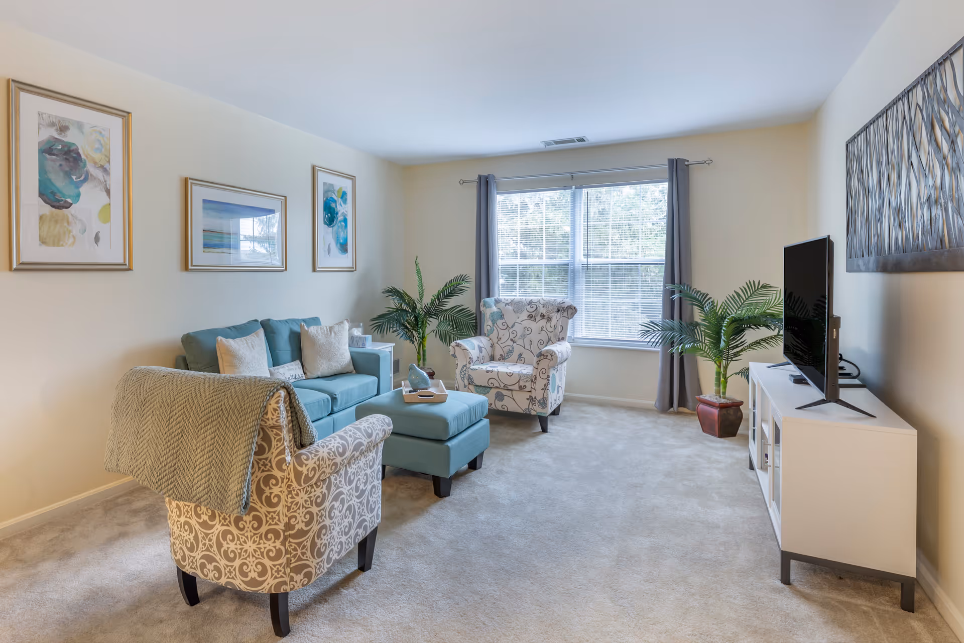 A bright living room with a large window covered by gray curtains. The room features a teal sofa with white pillows, two patterned armchairs, a matching teal ottoman, a white TV stand with a flat-screen TV, two potted plants, and three framed artworks on the walls. The floor is carpeted in a light beige color.