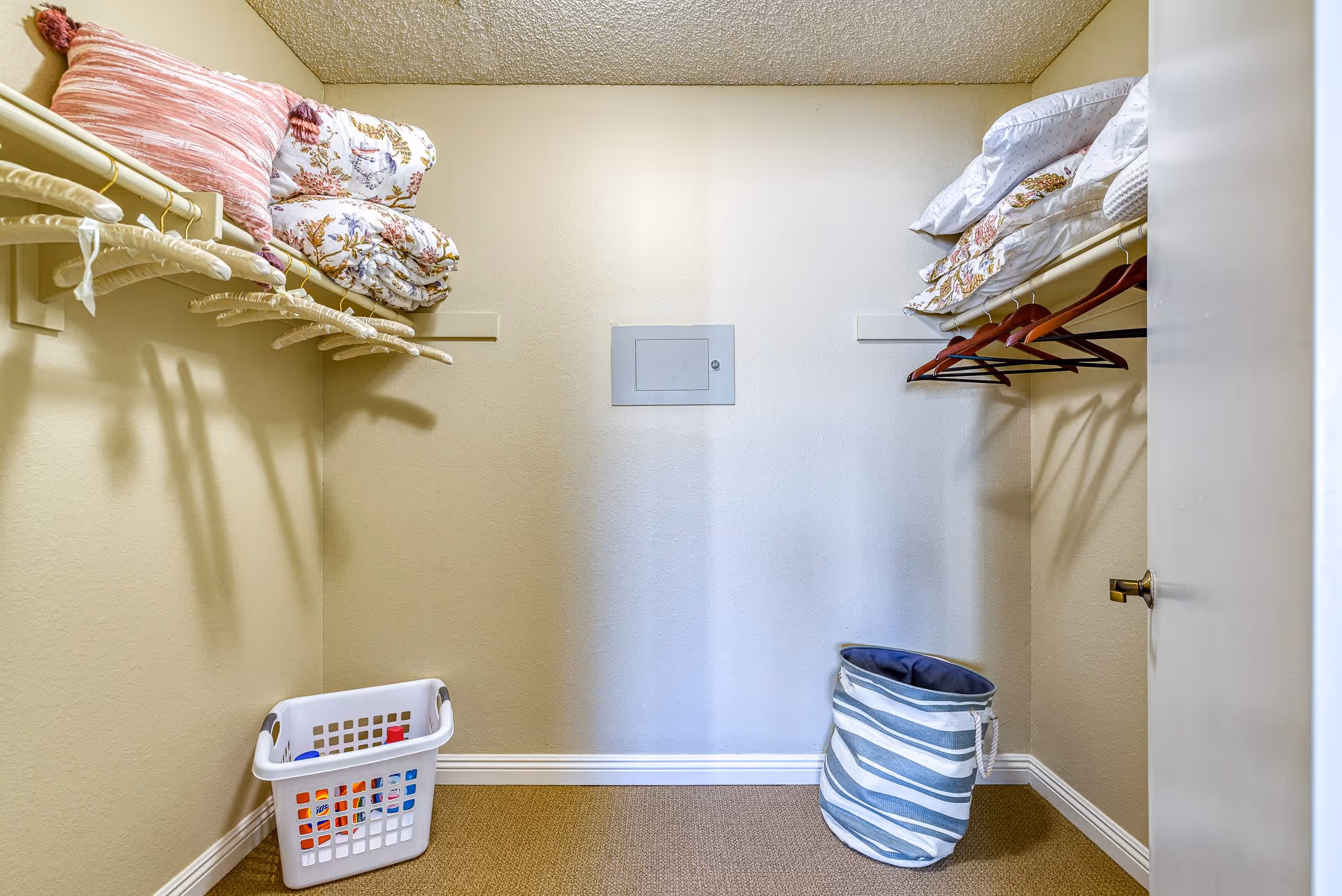 A small walk-in closet with beige walls and carpeted floor. There are shelves on both sides holding folded blankets and pillows. Several empty hangers are hanging on rods beneath the shelves. On the floor, there is a white laundry basket with some items inside and a blue and white striped fabric hamper.