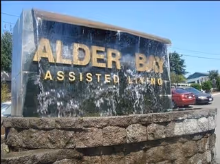 A stone and water feature sign for Alder Bay Assisted Living with water cascading down the dark stone surface and gold lettering. There are cars and houses visible in the background under a clear blue sky.