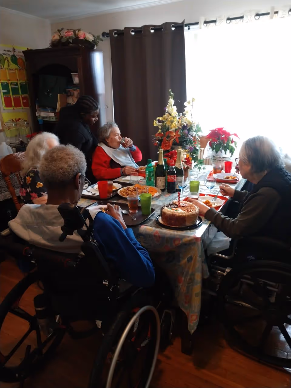 A group of elderly people seated around a dining table enjoying a meal together. The table is set with plates of food, drinks, and a cake with a single candle. There are flower arrangements on the table and a caregiver standing behind one of the seated individuals. The room has a cabinet and a window with curtains in the background.