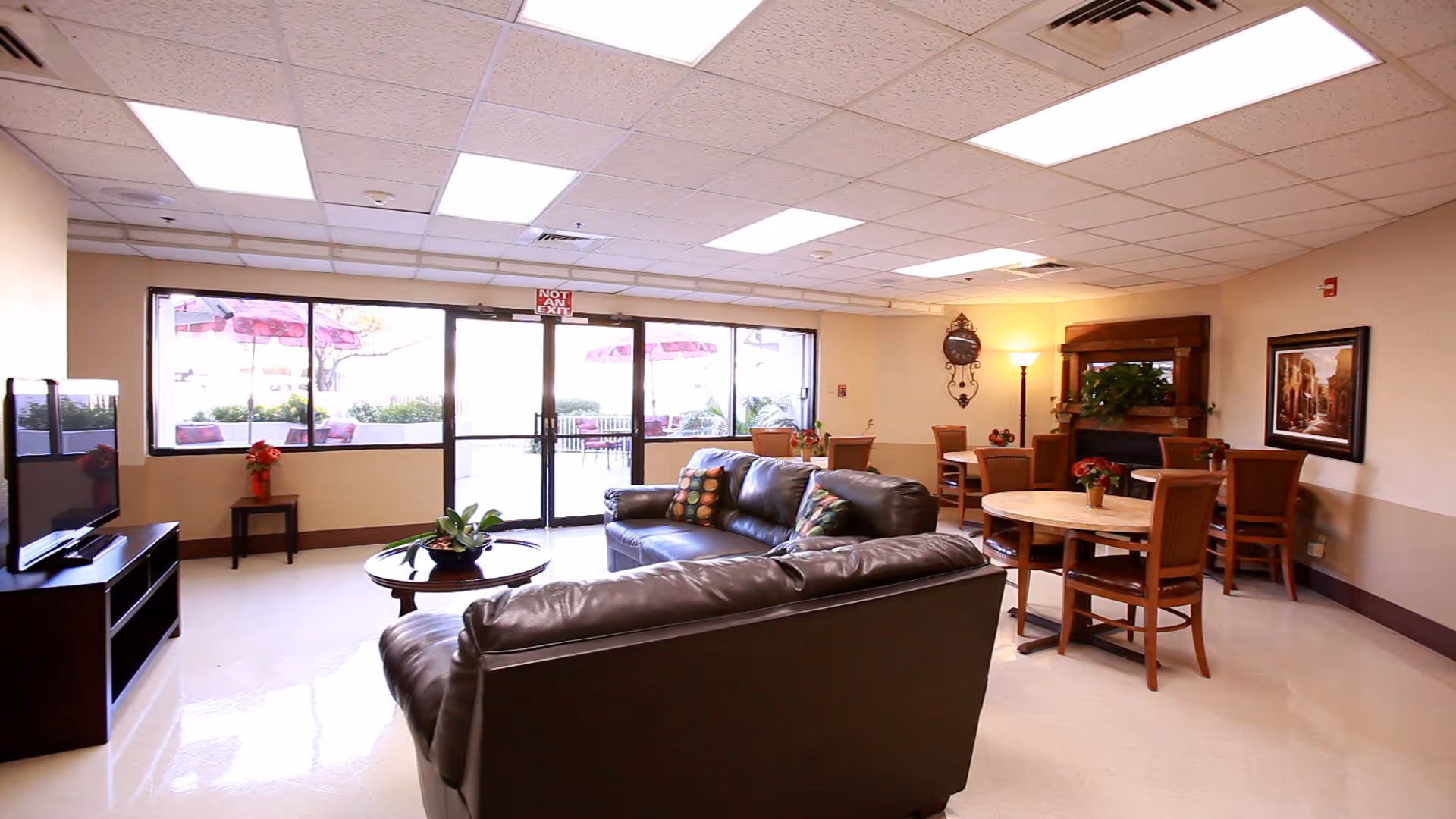 A bright and spacious living room area with large windows and glass doors leading to an outdoor patio. The room features two dark leather sofas with colorful pillows, a round wooden coffee table with a plant, a flat-screen TV on a stand, several wooden dining tables with chairs, a floor lamp, a decorative wall clock, and framed artwork on the walls.