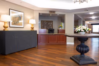 A spacious reception area with a wooden floor, a dark wooden front desk with a computer and flowers on it, a round wooden table with a flower arrangement in the foreground, two table lamps on a sideboard against the left wall, and framed artwork above the sideboard.